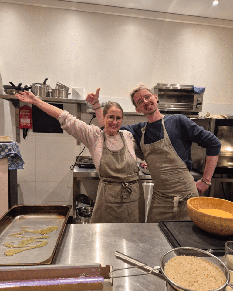 Magali, fondatrice du café La Tasse à Uccle, souriante en cuisine aux côtés d’un collègue, tous deux en tablier