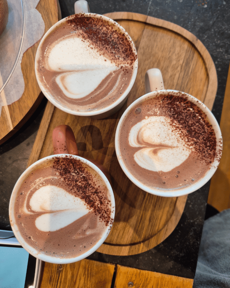 Trois mugs de chocolat chaud avec mousse et poudre de cacao, servis sur un plateau en bois