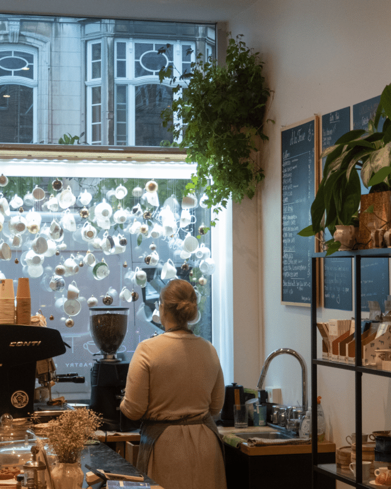 Vue de dos de Magali, fondatrice de La Tasse, en service derrière le comptoir du café à Uccle, avec tasses suspendues à la vitrine