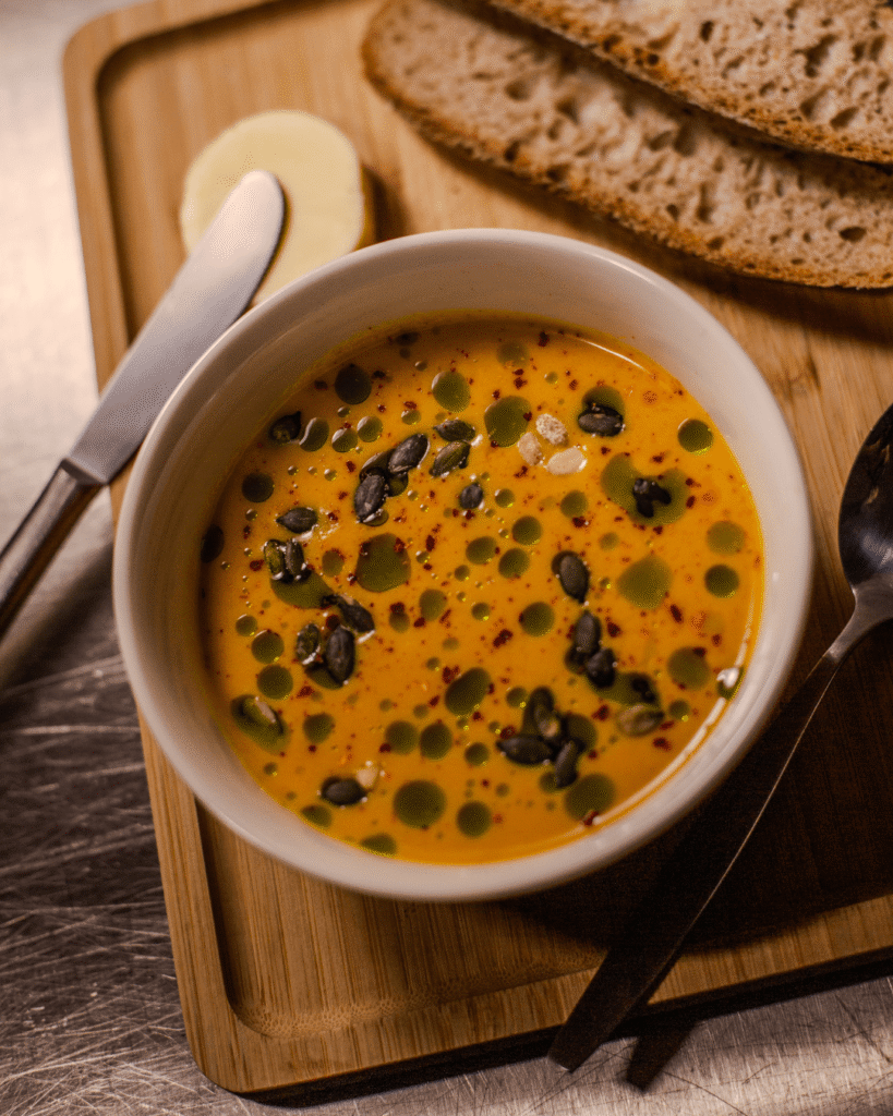 Bol de soupe colorée à l’huile d’herbes et graines, servi avec pain au levain et beurre à La Tasse