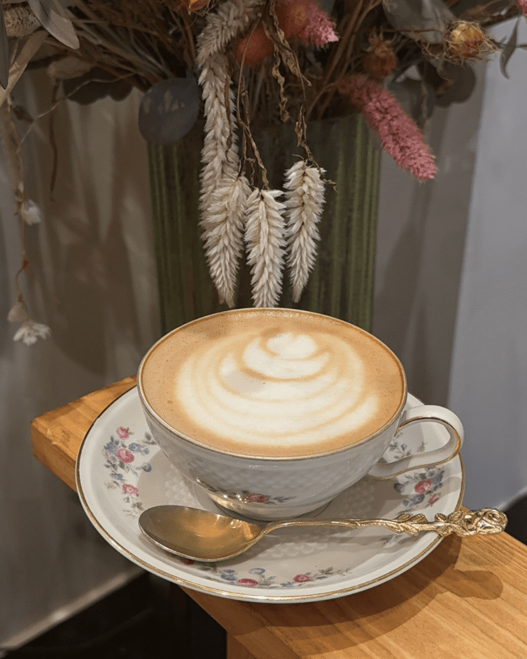 Tasse de latte posée sur une soucoupe fleurie avec bouquet sec en arrière-plan, ambiance cosy chez La Tasse