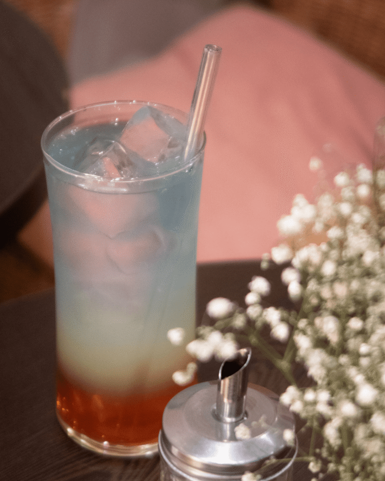 Boisson glacée colorée servie dans un grand verre avec paille en verre, posée sur table en bois foncé