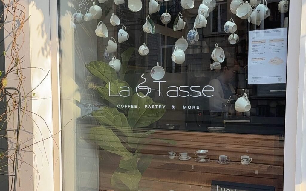 Devanture du café La Tasse à Uccle, avec logo visible sur la vitre et tasses suspendues en décoration intérieure