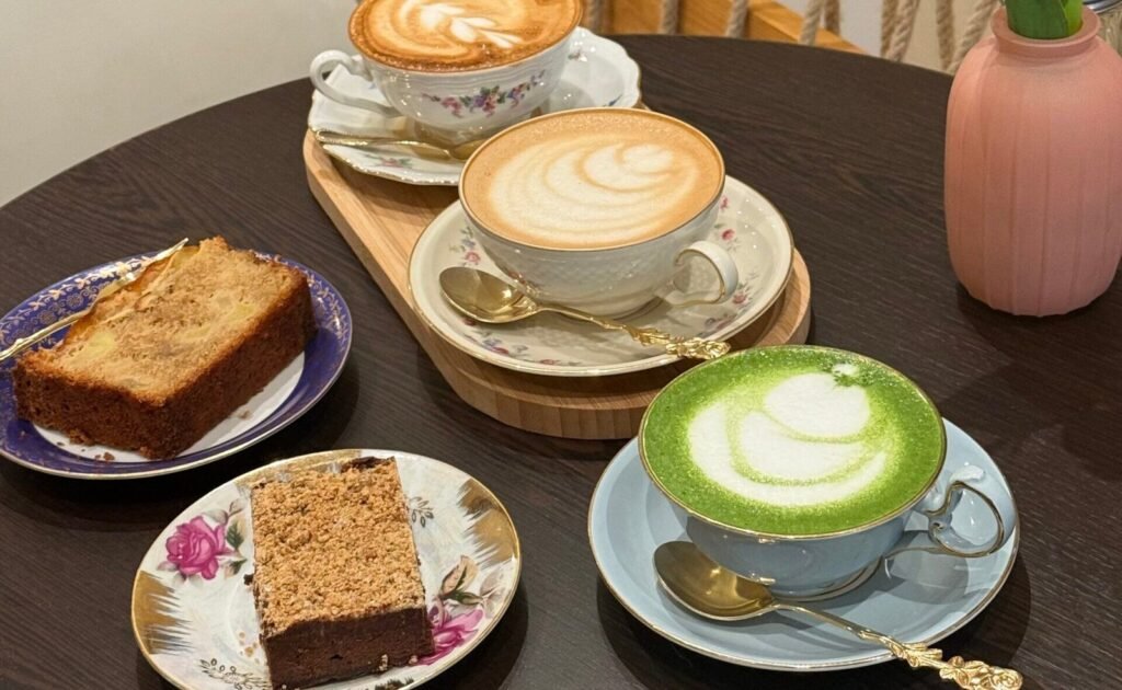 Trois boissons chaudes servies dans des tasses vintage accompagnées de deux parts de gâteau maison, sur une table du salon de thé La Tasse à Uccle